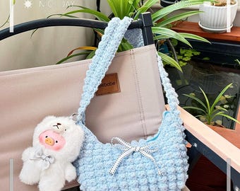 May include: A light blue, crocheted handbag with a matching strap and a small white bow. A white plush bear keychain with a pink face and a bow is attached to the bag. The text "Nice Day" is visible in the background.