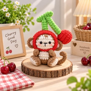 May include: A crocheted monkey amigurumi wearing a red cherry-shaped hood with a green stem and leaf. The monkey has brown and white yarn details, black eyes, and a stitched mouth. It sits on a round wooden slice, surrounded by cherries.