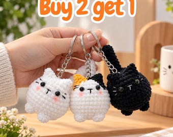 May include: Three round, crocheted cat keychains with metal rings. One is solid white, one is white with black and orange patches, and one is solid black. Each has black bead eyes, embroidered whiskers, and pink cheeks. Text reads: Buy 2 get 1.