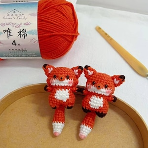 May include: Two small, handmade crochet foxes with orange and white fur, black ears, and pink cheeks. A ball of orange yarn and a white yarn ball are in the background, along with a crochet hook. The text on the yarn label reads "Susan's Family".