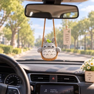 以下が含まれることがあります： 車のバックミラーに茶色の紐で吊るされた、グレーと白の編みぐるみのキャラクター。近くには「Good Drive Good Mood」という文字と葉のアイコンが刻まれた長方形の木製タグが下がっている。ダッシュボードには小さなデイジーの鉢植えが置かれている。