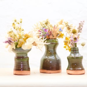 Puede incluir: Tres pequeños jarrones de cerámica en tonos verdes y marrones, cada uno lleno de flores secas. Los jarrones tienen un aspecto rústico y hecho a mano y están dispuestos sobre una superficie blanca.