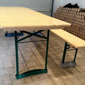 Könnte beinhalten: Ein hellfarbener Picknicktisch aus Holz mit passender Bank. Der Tisch hat eine rechteckige Platte und grüne Metall-Klappbeine. Die Bank hat ein ähnliches Design. Der Tisch ist etwa 244 cm lang.