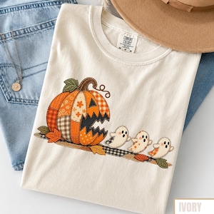 May include: An ivory t-shirt featuring a large, patchwork-style jack-o'-lantern pumpkin graphic with orange and brown patterns, accompanied by three small, white ghost figures. The shirt has a Comfort Colors label at the inner neckline.