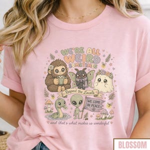 May include: Pink t-shirt with a whimsical graphic featuring a Bigfoot, a moth, a ghost, a dinosaur, and an alien. The text reads "WE'RE ALL WEIRD SOMETIMES" and "Respect the Locals". The shirt also has the text "WE COME IN PEACE" and "and that's what makes us wonderful".