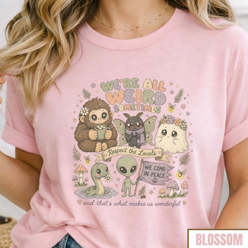 May include: Pink t-shirt with a whimsical graphic featuring a Bigfoot, a moth, a ghost, a dinosaur, and an alien. The text reads "WE'RE ALL WEIRD SOMETIMES" and "Respect the Locals". The shirt also has the text "WE COME IN PEACE" and "and that's what makes us wonderful".