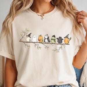 Peut inclure: Un t-shirt couleur cr&egrave;me arborant une illustration graphique de huit petits oiseaux perch&eacute;s sur une branche. Les oiseaux portent divers costumes, dont un chapeau de sorci&egrave;re, un drap de fant&ocirc;me, une citrouille, un smoking, une &eacute;charpe et des ailes.
