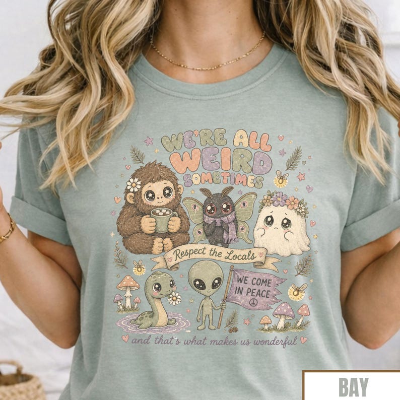 May include: A light blue t-shirt with a whimsical graphic. The design features a Bigfoot character, a fairy, a ghost, an alien, and a dinosaur. Text includes "We're all weird sometimes", "Respect the Locals", and "We come in peace".