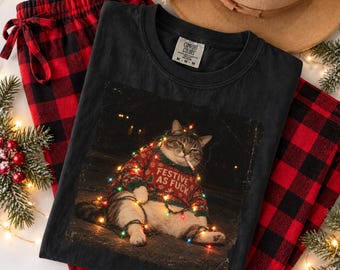 May include: A black crewneck t-shirt featuring a graphic of a cat wearing a red patterned sweater with the text FESTIVE AS FUCK, wrapped in colorful Christmas lights. The shirt is paired with red and black buffalo plaid pajama pants.