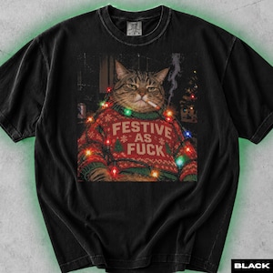 Peut inclure: Un t-shirt noir à manches courtes avec un imprimé représentant un chat tigré portant un pull de Noël rouge et vert avec le texte FESTIVE AS FUCK. Le chat est représenté avec une cigarette et entouré de guirlandes lumineuses colorées.