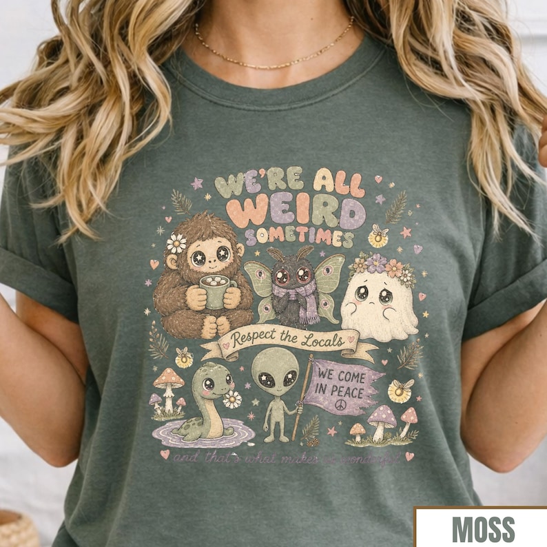 May include: Moss green t-shirt with a whimsical graphic. The design includes a Bigfoot holding a mug, a moth with a scarf, a ghost with a flower crown, a Loch Ness Monster, and an alien holding a flag that says "We Come in Peace". The text "We're All Weird Sometimes" is also featured.