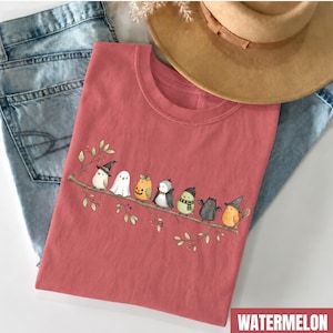 Peut inclure: Un t-shirt couleur past&egrave;que orn&eacute; d'un motif repr&eacute;sentant sept petits oiseaux perch&eacute;s sur une branche. Les oiseaux portent divers costumes, dont une sorci&egrave;re, un fant&ocirc;me, une citrouille, un vampire, un oiseau avec une &eacute;charpe, une chauve-souris.