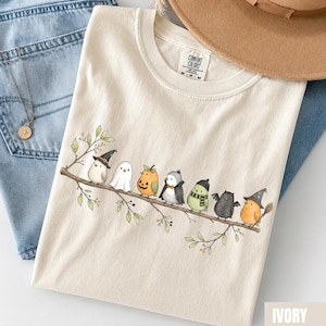 Peut inclure: Un t-shirt ivoire avec une illustration de sept petits oiseaux perch&eacute;s sur une branche, chacun portant un costume d'Halloween diff&eacute;rent, incluant un chapeau de sorci&egrave;re, un drap de fant&ocirc;me, une citrouille, une cape, une &eacute;charpe et des ailes.