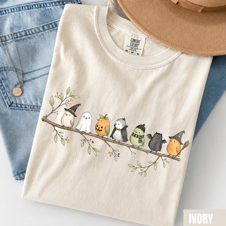 Peut inclure: Un t-shirt ivoire avec une illustration de sept petits oiseaux perch&eacute;s sur une branche, chacun portant un costume d'Halloween diff&eacute;rent, incluant un chapeau de sorci&egrave;re, un drap de fant&ocirc;me, une citrouille, une cape, une &eacute;charpe et des ailes.