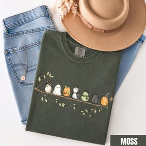 Peut inclure: Un t-shirt vert olive fonc&eacute; orn&eacute; d'un motif repr&eacute;sentant huit petits oiseaux perch&eacute;s sur une branche, chacun portant un costume d'Halloween diff&eacute;rent, dont un chapeau de sorci&egrave;re, un drap de fant&ocirc;me, une citrouille et une &eacute;charpe, avec un jean bleu.