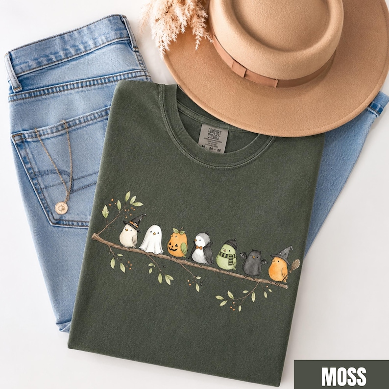 Peut inclure: Un t-shirt vert olive fonc&eacute; orn&eacute; d'un motif repr&eacute;sentant huit petits oiseaux perch&eacute;s sur une branche, chacun portant un costume d'Halloween diff&eacute;rent, dont un chapeau de sorci&egrave;re, un drap de fant&ocirc;me, une citrouille et une &eacute;charpe, avec un jean bleu.