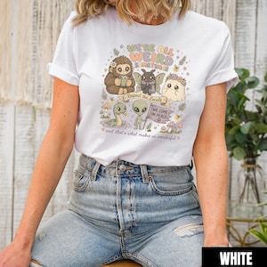 May include: White t-shirt featuring a whimsical illustration with the text "WE'RE ALL WEIRD SOMETIMES". The design includes various cartoon creatures, mushrooms, and the phrases "Respect the Locals" and "WE COME IN PEACE".