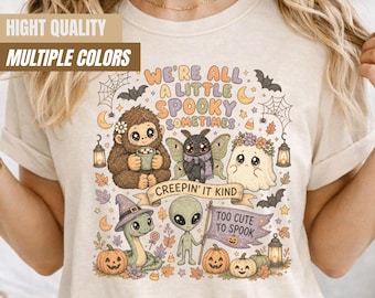 May include: An ivory t-shirt featuring a colorful graphic print with the text "WE'RE ALL A LITTLE SPOOKY SOMETIMES", "CREEPIN' IT KIND", and "TOO CUTE TO SPOOK". The design includes a sasquatch, a moth, a ghost, a snake, and an alien with pumpkins.