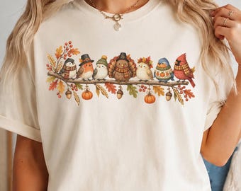 May include: A cream-colored t-shirt featuring a graphic of seven birds perched on a branch. The birds wear hats and scarves, surrounded by autumn leaves, acorns, and pumpkins. The design includes a turkey, a cardinal, and other small birds.