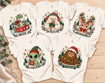 May include: Five cream-colored t-shirts featuring holiday-themed illustrations of mythical creatures. The designs include Nessie in a mug, a Yeti with a candy cane, a Jackalope with antlers, an Alien with an ornament, and Bigfoot with lights.