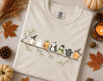 May include: A cream-colored t-shirt featuring a graphic of seven small birds perched on a branch, each dressed in a different Halloween costume including a witch, ghost, pumpkin, vampire, scarf, bat, and wizard, with the Comfort Colors brand label visible.