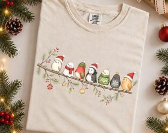 May include: An ivory-colored t-shirt featuring a graphic illustration of seven small birds perched on a pine branch. The birds are wearing various winter accessories including Santa hats, scarves, and sweaters, with small ornaments hanging from the branch.