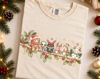Könnte beinhalten: Ein elfenbeinfarbenes T-Shirt mit einem Grafikdruck von einem Yeti, Alien, Motte, Reh, Dinosaurier und Lamm, die auf einem mit Stechpalmen, Tannenzapfen und Sternen geschmückten Ast sitzen. Jede Kreatur trägt einen Winterschal oder eine Mütze.