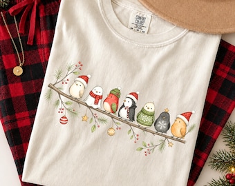 May include: An ivory t-shirt featuring a graphic of seven small birds perched on a branch. The birds wear winter accessories like Santa hats, scarves, and sweaters. The branch is decorated with pine needles, red berries, and hanging ornaments.