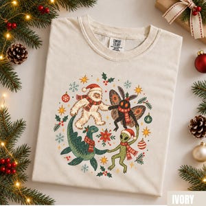 May include: An ivory t-shirt featuring a circular graphic of a yeti, a moth, a sea monster, and an alien holding hands. Each creature wears a red and white scarf and holiday hat. The design includes holly, snowflakes, stars, and ornaments.