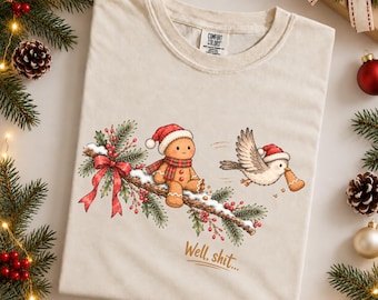 May include: An ivory t-shirt featuring a graphic of a gingerbread person wearing a Santa hat and scarf sitting on a snowy pine branch with a red bow. A bird wearing a Santa hat flies nearby holding a gingerbread cookie. The text reads, Well, shit...