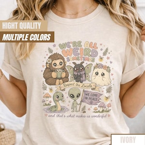 May include: Ivory-colored t-shirt featuring a whimsical illustration with the text "WE'RE ALL WEIRD SOMETIMES." The design includes a Bigfoot, a moth, a ghost, a dinosaur, and an alien. The phrase "Respect the Locals" is on a banner.