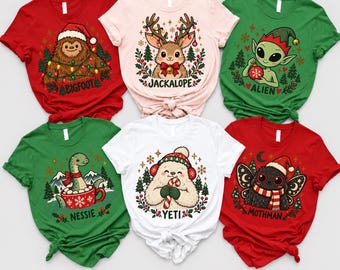 May include: Six short-sleeve t-shirts featuring Christmas-themed illustrations of Bigfoot, a jackalope, an alien, Nessie, a yeti, and Mothman. The shirts are red, light pink, green, and white, each displaying the creature's name in black text.