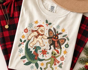 May include: An ivory t-shirt featuring a graphic print of a yeti, a moth, a sea monster, and an alien, all wearing red and white striped scarves and Santa hats. The design is surrounded by festive elements like snowflakes, stars, and holly.