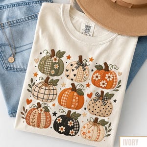 May include: An ivory short-sleeve t-shirt featuring a graphic print of nine pumpkins in various patterns including plaid, polka dots, and floral designs. The shirt has a Comfort Colors label at the neck and is decorated with small stars and hearts.