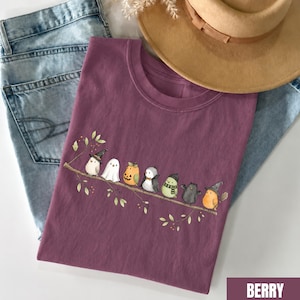 Peut inclure: Un t-shirt couleur baie avec une illustration graphique de sept petits oiseaux perch&eacute;s sur une branche. Les oiseaux portent divers costumes, dont un chapeau de sorci&egrave;re, un drap de fant&ocirc;me, une citrouille, un n&oelig;ud papillon, une &eacute;charpe et des ailes.
