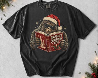 Könnte beinhalten: Ein anthrazitfarbenes Kurzarm-T-Shirt mit einer Grafik einer pelzigen Kreatur, die eine rote Weihnachtsmannmütze und eine schwarze Sonnenbrille trägt und ein rotes Buch mit dem Titel BIGFOOT'S CHRISTMAS LIST liest, verziert mit Schneeflocken.