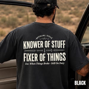 May include: Black t-shirt with the text "OFFICIAL TITLE KNOWER OF STUFF FIXER OF THINGS" in white. The shirt also has the text "Est. When Things Broke • Still On Duty" in white. The word "BLACK" is printed at the bottom right.