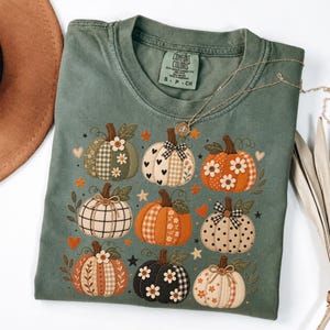 May include: A sage green short-sleeve t-shirt featuring a graphic print of nine pumpkins in various patterns, including plaid, polka dots, and floral designs. The shirt has a crew neckline and a Comfort Colors brand label printed on the inside collar.