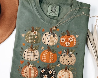 May include: A sage green short-sleeve t-shirt featuring a graphic print of nine pumpkins in various patterns, including plaid, polka dots, and floral designs. The pumpkins are decorated with small flowers, bows, hearts, and stars.