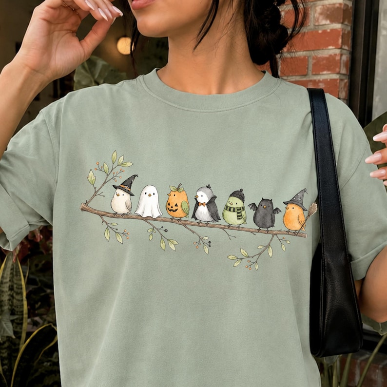 Peut inclure: Un t-shirt vert sauge avec une illustration graphique de sept petits oiseaux perch&eacute;s sur une fine branche. Les oiseaux portent divers costumes, dont un chapeau de sorci&egrave;re, un drap de fant&ocirc;me, une citrouille, un smoking, une &eacute;charpe, des ailes de chauve-souris et un chapeau.