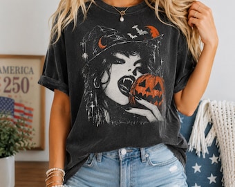 May include: A charcoal gray short-sleeve t-shirt featuring a graphic print of a witch wearing a hat, holding a jack-o'-lantern pumpkin. The design includes a spider, bats, and a crescent moon in orange and white ink on the front of the garment.