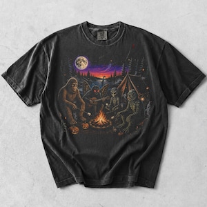 Peut inclure: Un t-shirt gris anthracite à manches courtes avec une impression graphique représentant Bigfoot, une créature ailée, un extraterrestre et un monstre des marais assis autour d'un feu de camp la nuit, sous une pleine lune, avec une tente.