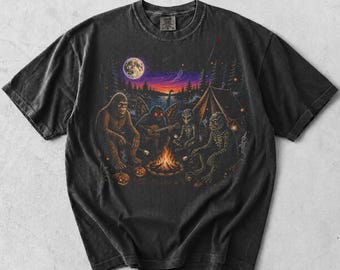 May include: A charcoal gray short-sleeve t-shirt featuring a graphic print of Bigfoot, a moth-like creature, an alien, and a swamp monster sitting around a campfire at night. The scene includes a full moon, a tent, and small pumpkins.