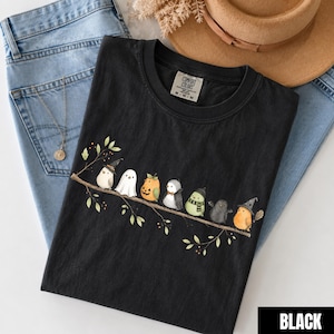 Peut inclure: Un t-shirt noir &agrave; col rond avec un motif repr&eacute;sentant sept petits oiseaux perch&eacute;s sur une branche. Les oiseaux portent divers costumes, notamment un chapeau de sorci&egrave;re, un drap de fant&ocirc;me, une citrouille, un n&oelig;ud papillon, une &eacute;charpe et des ailes.