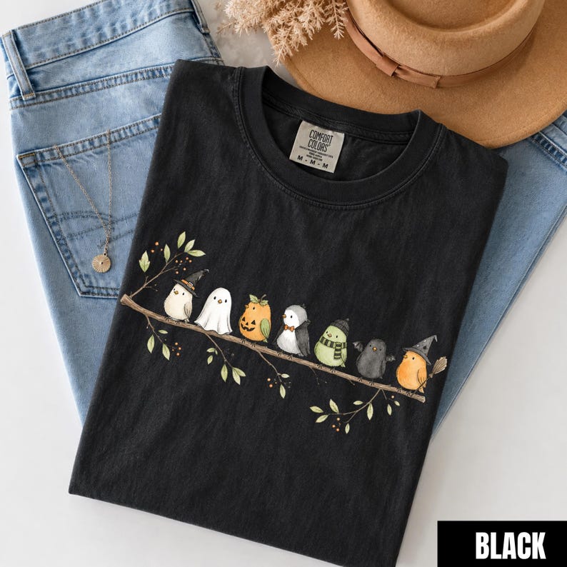Peut inclure: Un t-shirt noir &agrave; col rond avec un motif repr&eacute;sentant sept petits oiseaux perch&eacute;s sur une branche. Les oiseaux portent divers costumes, notamment un chapeau de sorci&egrave;re, un drap de fant&ocirc;me, une citrouille, un n&oelig;ud papillon, une &eacute;charpe et des ailes.
