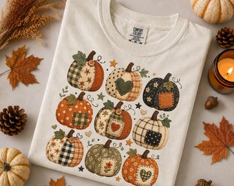 May include: A cream-colored t-shirt featuring a graphic print of nine pumpkins with various patchwork patterns, including plaid, polka dots, and floral designs, accented with small stars and hearts. The inner neck label reads Comfort Colors.