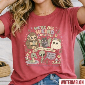 May include: A watermelon-colored t-shirt with a whimsical graphic. The design features a friendly Bigfoot, a moth, a ghost, an alien, and a snake, with the text "WE'RE ALL WEIRD SOMETIMES" and "Respect the Locals".