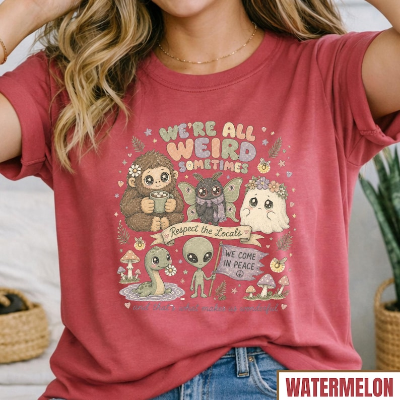 May include: A watermelon-colored t-shirt with a whimsical graphic. The design features a friendly Bigfoot, a moth, a ghost, an alien, and a snake, with the text "WE'RE ALL WEIRD SOMETIMES" and "Respect the Locals".