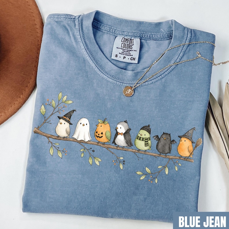 Peut inclure: Un t-shirt bleu &agrave; manches courtes orn&eacute; d'un motif repr&eacute;sentant sept oiseaux perch&eacute;s sur une branche, chacun portant un costume d'Halloween diff&eacute;rent, notamment une sorci&egrave;re, un fant&ocirc;me, une citrouille, un vampire et une chauve-souris.