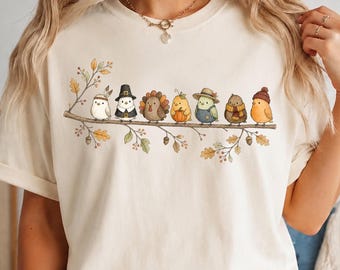 May include: A cream-colored short-sleeve t-shirt featuring a graphic of seven small birds perched on a branch. The birds are dressed in various autumn-themed outfits, including hats, scarves, and a turkey costume, surrounded by oak leaves and acorns.